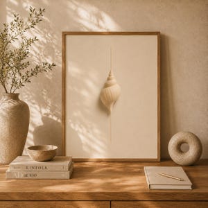 Könnte beinhalten: Ein gerahmter Kunstdruck mit einer zentrierten, hellbeigen spiralförmigen Muschelgrafik, die von einer dünnen vertikalen Goldlinie durchschnitten wird. Das Kunstwerk befindet sich in einem schlichten Holzrahmen mit neutraler Farbpalette.