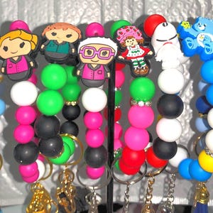 May include: Colorful beaded keychains with cartoon character charms. The keychains feature various bead colors, including pink, green, black, and blue. Charms include cartoon characters like Strawberry Shortcake and the Care Bear.