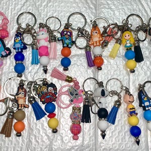 May include: Collection of keychains with various cartoon character charms, including Snoopy, Charlie Brown, and Bluey. Each keychain features colorful beads, tassels, and a metal keyring. Some have fluffy pom-poms.