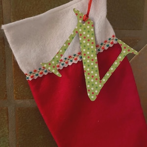 May include: A red Christmas stocking with a white cuff and a green letter "N" ornament. The letter is decorated with red and white polka dots and hangs from a red ribbon. The stocking is trimmed with a decorative border.