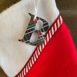 May include: A Christmas stocking with a white felt top and a red felt bottom, trimmed with white and red rope. A wooden letter "B" with a plaid pattern hangs from a white ribbon. The plaid includes green, red, black, and brown.
