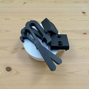 Puede incluir: Una colección de objetos negros de forma geométrica, que incluyen soportes y piezas curvas, están dispuestos sobre una plataforma circular blanca. Los artículos están hechos de un material mate y sobre un fondo claro de vetas de madera.