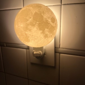 May include: A white moon-shaped night light plugged into an electrical outlet. The light is on and illuminating the surrounding area.