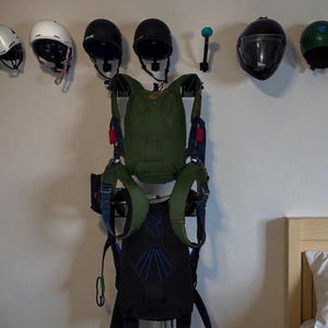 Puede incluir: Una exhibición mural de cascos y un arnés de paracaídas. Los cascos son de varios colores, incluyendo blanco, negro y verde. El arnés de paracaídas es principalmente verde y negro, con detalles azules y rojos. Los artículos están dispuestos contra una pared blanca.