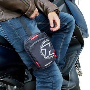 May include: Black leg bag with a white logo and red accents, worn on the thigh. The bag is attached to a black strap and is being worn by a person wearing blue jeans and a brown leather jacket. The bag has the word "Motocard" printed on it.