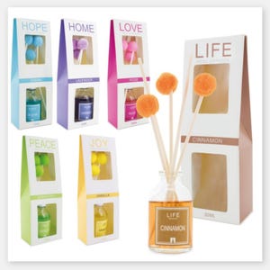 Puede incluir: Una colección de difusores de varillas con varios aromas, incluyendo Ocean, Lavanda, Rosa y Canela. Cada difusor presenta una botella de vidrio con líquido coloreado y un conjunto de varillas de caña rematadas con un pompón decorativo. El embalaje incluye las palabras "HOPE", "HOME", "LOVE", "PEACE", "JOY" y "LIFE".