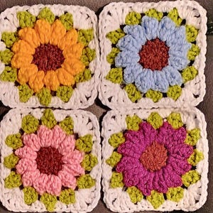 Puede incluir: Cuatro cuadrados de ganchillo con diseños de flores. Cada cuadrado presenta una flor de diferente color: amarillo, azul, rosa y morado. Las flores tienen centros marrones y hojas verdes, sobre un fondo blanco.