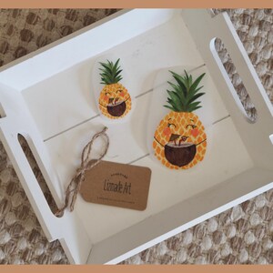 Könnte beinhalten: Ein weißes Holztablett mit zwei Ananas-Aufklebern und einem kleinen Etikett. Die Aufkleber zeigen lächelnde Ananas mit einem tropischen Getränk. Auf dem Etikett steht "Lizmade Art". Das Tablett hat ausgeschnittene Griffe.