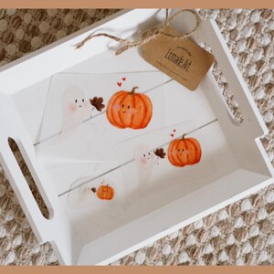 Könnte beinhalten: Ein weißes Tablett mit drei handgemalten Illustrationen von Geistern und Kürbissen. Die Illustrationen sind in einem skurrilen Stil mit einfachen Linien und leuchtenden Farben gehalten. Das Tablett ist mit einem kleinen Etikett verziert, auf dem "Limeade Art" steht.