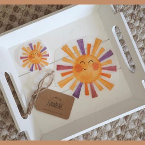 Könnte beinhalten: Ein weißes Holztablett mit Griffen enthält zwei sonnenförmige Aufkleber mit orangefarbenen, lila und gelben Strahlen. Ein kleines Etikett mit dem Text "Lizmade Art" ist mit Bindfaden gebunden. Das Tablett steht auf einer strukturierten, neutralfarbenen Oberfläche.