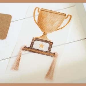 Könnte beinhalten: Aquarell-Illustration eines goldenen Trophäen-Aufklebers mit einem Stern auf weißem Hintergrund. Der Aufkleber hat einen braunen Rand und eine braune Quaste. Ein braunes Etikett mit dem Wort "Art" ist sichtbar.