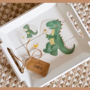 Könnte beinhalten: Ein weißes Holztablett mit zwei grünen Dinosaurier-Aufklebern und einem gelben Küken-Aufkleber. Das Tablett hat auch ein braunes Etikett mit dem Text "Lemonade Art" darauf.