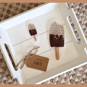 Könnte beinhalten: Ein weißes Holztablett mit zwei Aufklebern in Eisform. Die Aufkleber haben eine braune Schokoladenbeschichtung mit Streuseln und einen Holzstiel. Ein braunes Etikett mit dem Text "Lizmade Art" ist mit Bindfaden befestigt.