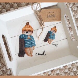 Könnte beinhalten: Zwei weiße Holztabletts mit einem braunen und orangefarbenen Bären, der einen blau-weiß gestreiften Pullover und einen braunen Hut trägt und ein braunes Surfbrett hält. Der Bär befindet sich auf einem weißen Aufkleber mit dem Wort "cool" in Schwarz. Das kleinere Tablett hat einen ähnlichen Aufkleber mit einem kleineren Bären und dem Wort "cool" in Schwarz.
