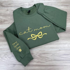 May include: A green sweatshirt with the text "cat mom" in gold lettering and a gold bow. The left sleeve has the names "Remy, Birdie, Shay" and a paw print in gold lettering.