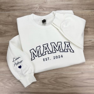 May include: White sweatshirt with the text "MAMA EST. 2024" in navy blue. The left sleeve has the text "Liam Asher" in navy blue with a small heart.
