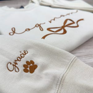 May include: A white sweatshirt with the text "dog mom" embroidered in brown on the front. The sweatshirt also has a brown embroidered bow and a brown paw print on the sleeve. The text "Gracie" is also embroidered in brown on the sleeve.