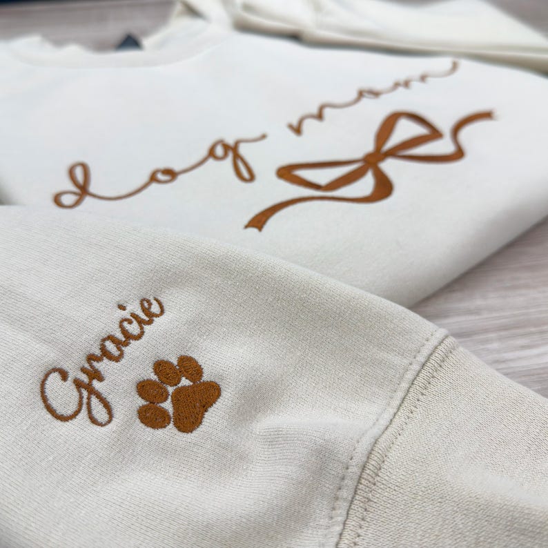 May include: A white sweatshirt with the text "dog mom" embroidered in brown on the front. The sweatshirt also has a brown embroidered bow and a brown paw print on the sleeve. The text "Gracie" is also embroidered in brown on the sleeve.