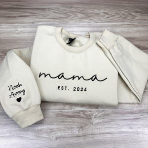 May include: A beige sweatshirt with the text "mama est. 2024" on the front. The left sleeve has the text "Noah Avery" and a small heart.
