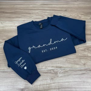 May include: A navy blue sweatshirt with the text "grandma est. 2024" on the front. The left sleeve has the names "Grayson, Isabella, Eliana" and a heart.
