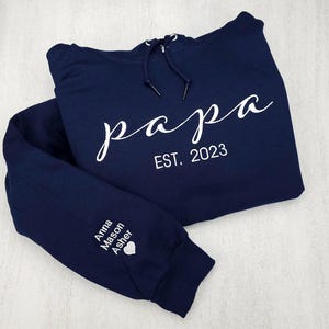 May include: A navy blue hoodie with the word "papa" in white script, and "EST. 2023" below it. The left sleeve has the names "Anna Mason Asher" and a heart symbol. The hoodie has a drawstring hood.