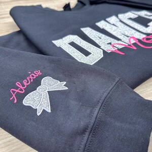 May include: Black sweatshirt with silver glitter lettering that says "Dance Mom" and a pink glitter bow on the sleeve with the name "Alexis" embroidered in pink.