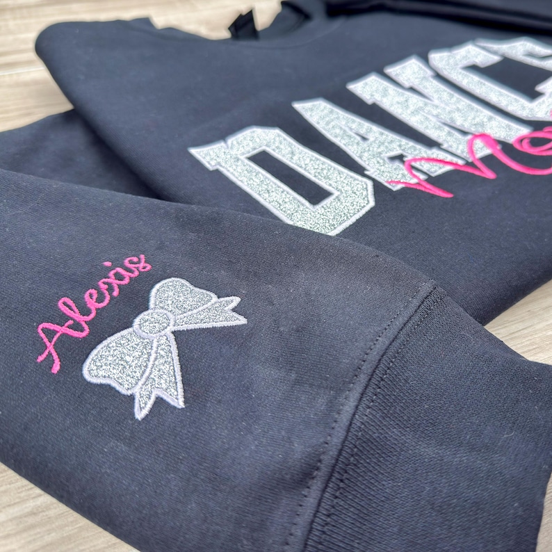 May include: Black sweatshirt with silver glitter lettering that says "Dance Mom" and a pink glitter bow on the sleeve with the name "Alexis" embroidered in pink.