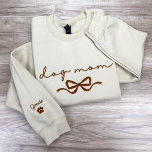 Sand sweatshirt folded neatly on a wooden surface, featuring dog mom; embroidery in brown script stitching across the design. With a brown bow on the chest underneath, and pets names in cursive on the sleeve with a paw print.