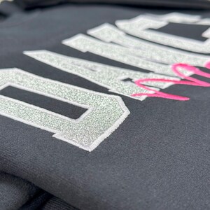 May include: Black sweatshirt with silver glitter lettering that reads "Dance Mom". The "Dance" is in large letters and the "Mom" is in smaller letters and pink.