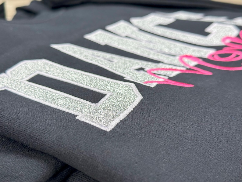 May include: Black sweatshirt with silver glitter lettering that reads "Dance Mom". The "Dance" is in large letters and the "Mom" is in smaller letters and pink.