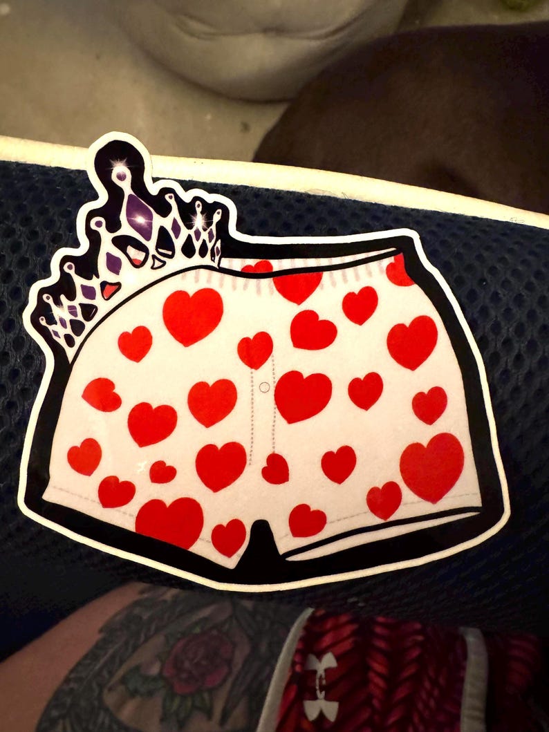 May include: A sticker featuring a pair of white underwear adorned with red hearts. A black and white crown is at the top. The underwear has a black border and a dotted line down the center.