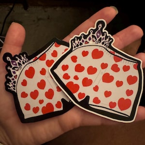 May include: Two stickers featuring a cartoon design of white underwear with red hearts and a crown. The stickers have a black outline and are held in a hand.