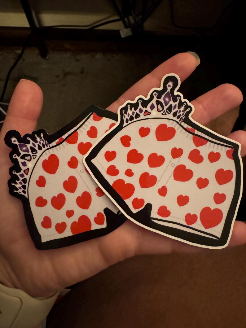 May include: Two stickers featuring a cartoon design of white underwear with red hearts and a crown. The stickers have a black outline and are held in a hand.