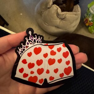 May include: A sticker in the shape of underwear with a crown design. The underwear is white with red hearts and a black outline. The crown is purple and white.