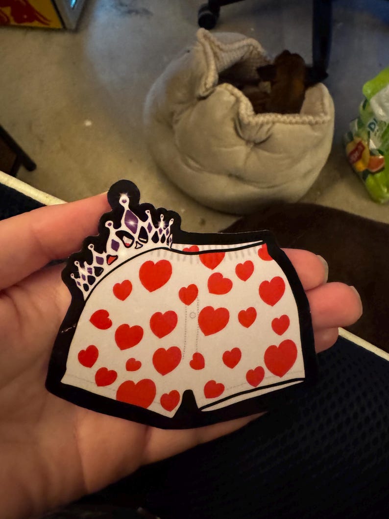 May include: A sticker in the shape of underwear with a crown design. The underwear is white with red hearts and a black outline. The crown is purple and white.