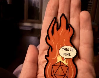 May include: A flame-shaped sticker with a D20 die and the text "THIS IS FINE." The sticker is primarily orange and red, with a black outline. The die is white with a black outline and the number 1.