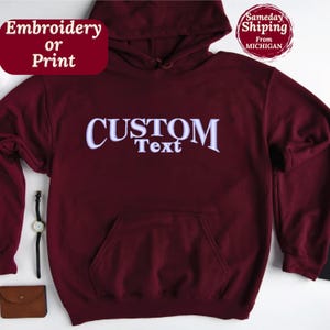 以下が含まれることがあります： ワインレッドのフード付きスウェットシャツに、薄いパープルの「CUSTOM Text」の文字が入っています。スウェットシャツは柔らかく快適な素材で作られており、カンガルーポケットが付いています。