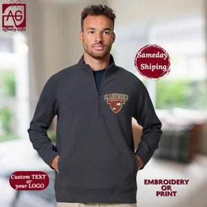 May include: A charcoal gray quarter-zip pullover with a small embroidered emblem. The emblem reads "ST. LAWRENCE" above a crest. The pullover has a front pocket on each side. The image also includes the text "Custom TEXT or your LOGO" and "EMBROIDERY OR PRINT".