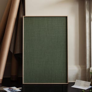 May include: A framed artwork with a textured, olive green fabric insert and a light wood frame. Rolls of brown paper and a dark fabric lean against the wall. Papers and a pencil are on the floor. The artwork is rectangular.