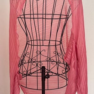 May include: A pink pleated scarf draped over a black metal dress form. The scarf has a flowing, lightweight appearance and is a vibrant shade of pink. The dress form is a decorative piece.