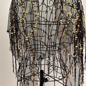 May include: Black sheer shawl with gold sequins and beaded fringe. The shawl is draped over a black wire dress form, showcasing its delicate design. The gold sequins are arranged in a cascading pattern.