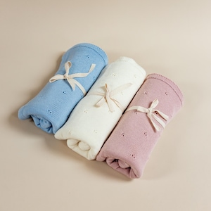 May include: Three rolled knit blankets in light blue, cream, and dusty rose. Each blanket features a delicate pointelle pattern of small eyelet holes and is secured with a thin, light beige ribbon tied in a bow around the center of the roll.