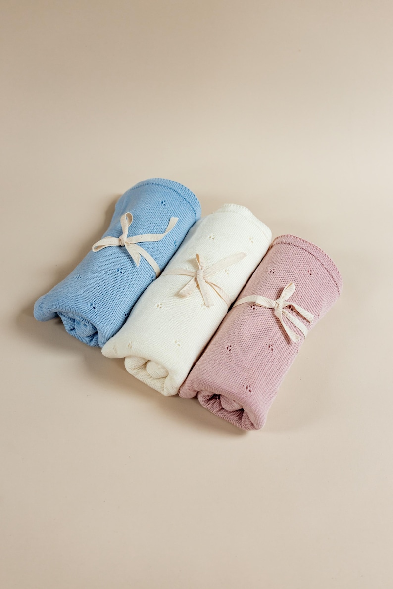 May include: Three rolled knit blankets in light blue, cream, and dusty rose. Each blanket features a delicate pointelle pattern of small eyelet holes and is secured with a thin, light beige ribbon tied in a bow around the center of the roll.