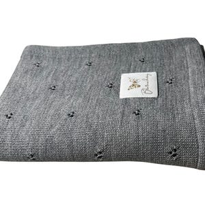 May include: A folded grey knit blanket featuring a repeating pattern of small, openwork eyelet holes. A rectangular white fabric label is sewn onto the top surface, displaying a gold butterfly graphic and the cursive text Blanchechou.