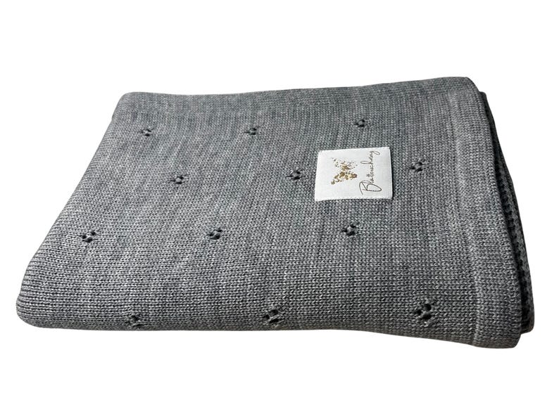 May include: A folded grey knit blanket featuring a repeating pattern of small, openwork eyelet holes. A rectangular white fabric label is sewn onto the top surface, displaying a gold butterfly graphic and the cursive text Blanchechou.