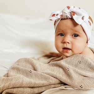 May include: A white ribbed baby headband featuring a knotted bow detail and a repeating pattern of small red cherries and brown stems, paired with a beige knit blanket that has a delicate pointelle texture with small eyelet hole details.