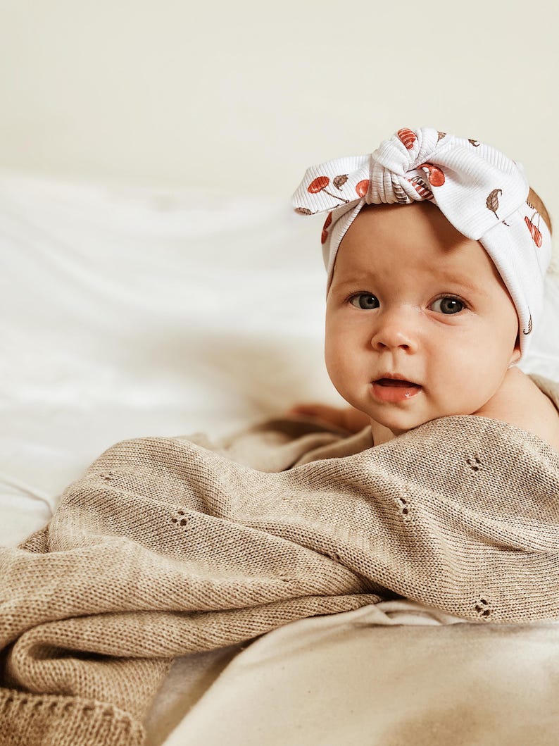May include: A white ribbed baby headband featuring a knotted bow detail and a repeating pattern of small red cherries and brown stems, paired with a beige knit blanket that has a delicate pointelle texture with small eyelet hole details.