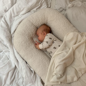 May include: A cream-colored, textured sherpa baby lounger with a raised, padded border. A baby is sleeping inside, wearing a long-sleeved white onesie with a small brown bear print. A soft, off-white knit blanket with a subtle dot pattern covers the baby.