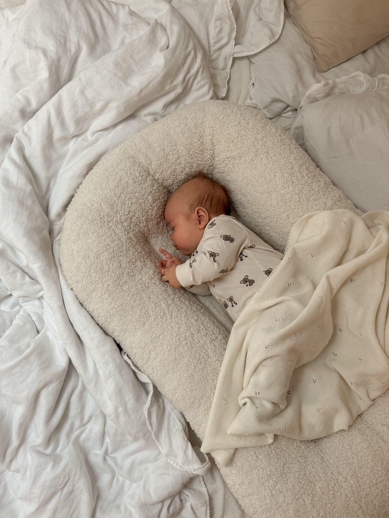 May include: A cream-colored, textured sherpa baby lounger with a raised, padded border. A baby is sleeping inside, wearing a long-sleeved white onesie with a small brown bear print. A soft, off-white knit blanket with a subtle dot pattern covers the baby.