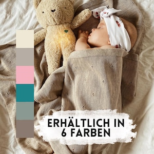 May include: A beige knitted baby blanket with a delicate eyelet pattern, a plush teddy bear, and a baby wearing a white headband with a cherry print. A vertical color palette shows six swatches, and text reads: ERH&Auml;LTLICH IN 6 FARBEN.